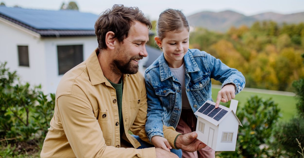 Little girl with her father holding paper model of house with the solar panels, explaining how it works.Alternative energy, saving resources and sustainable lifestyle concept.